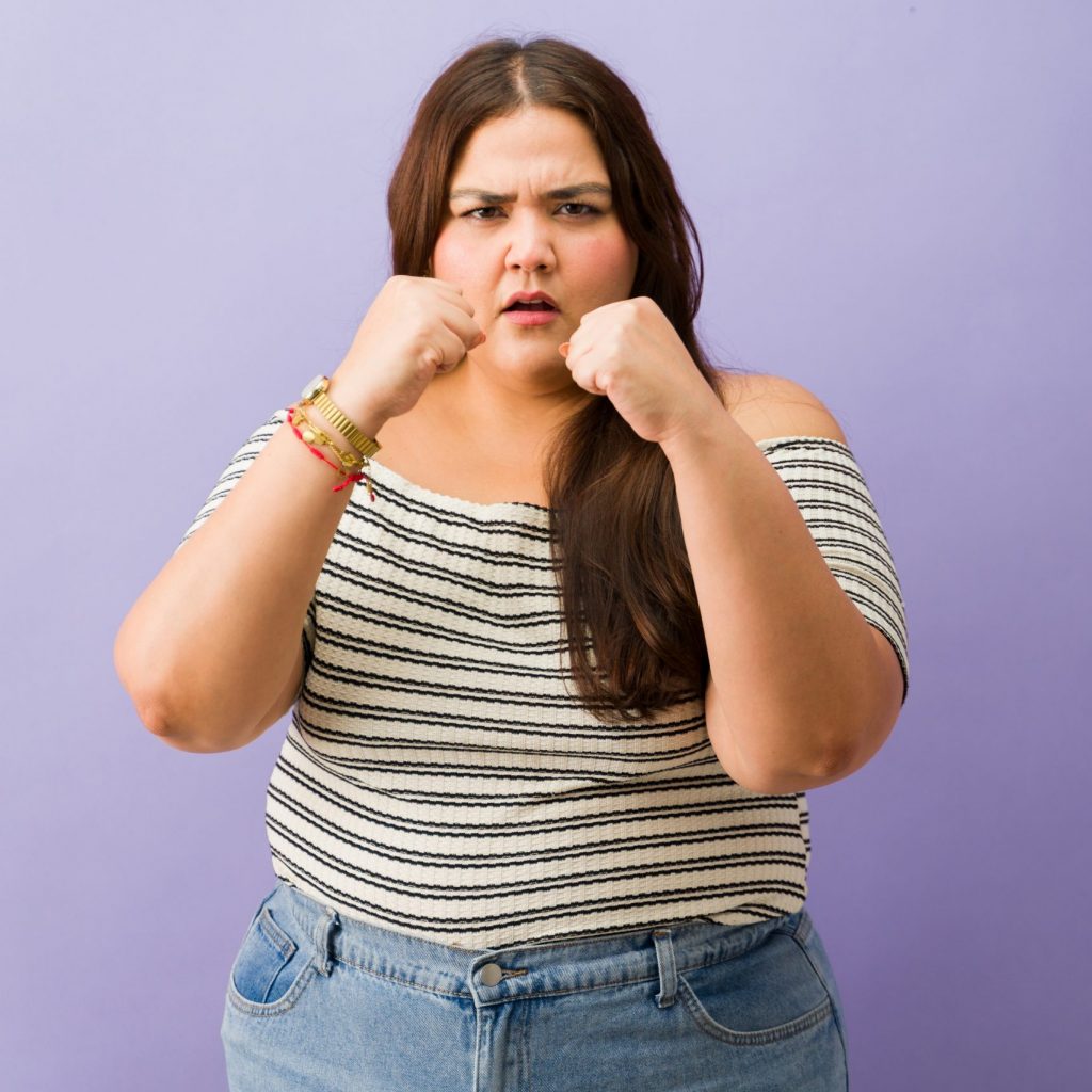 Selbstbewusste Frau mit geballten Fäusten vor lila Hintergrund, die Stärke und Empowerment für die Frauenkurse symbolisiert.