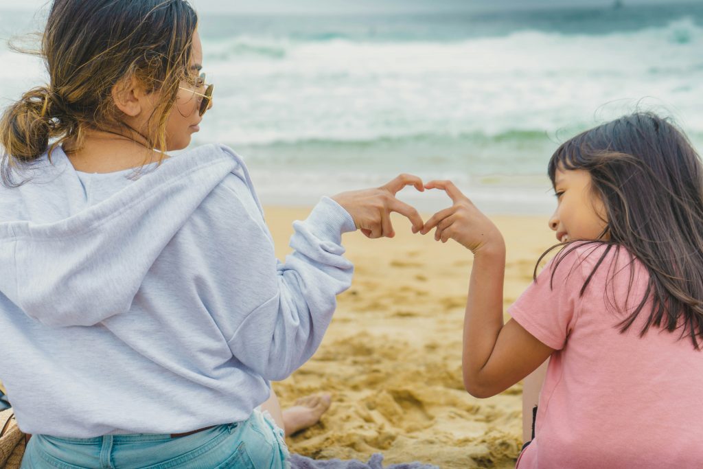 Mutter und Tochter am Strand formen ein Herz mit ihren Händen, was die Verbindung und Freude der Mutter-Tochter-Kurse symbolisiert.