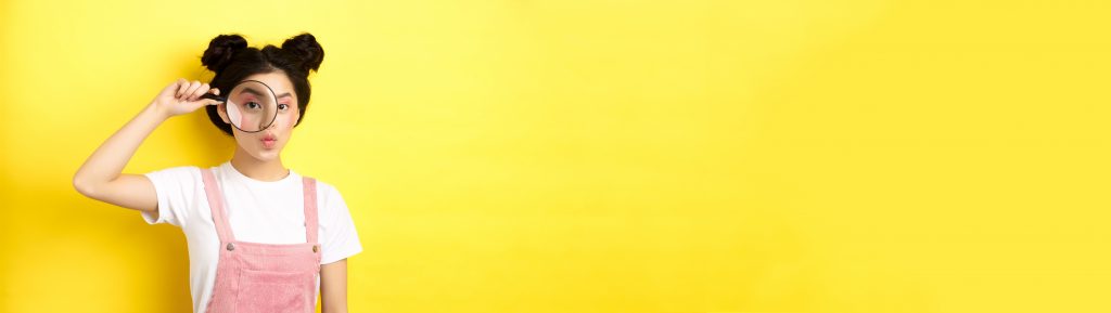 Junge Frau hält eine Lupe vor ihr Gesicht vor gelbem Hintergrund. Das Bild steht für Entdeckungsfreude und Fragen.