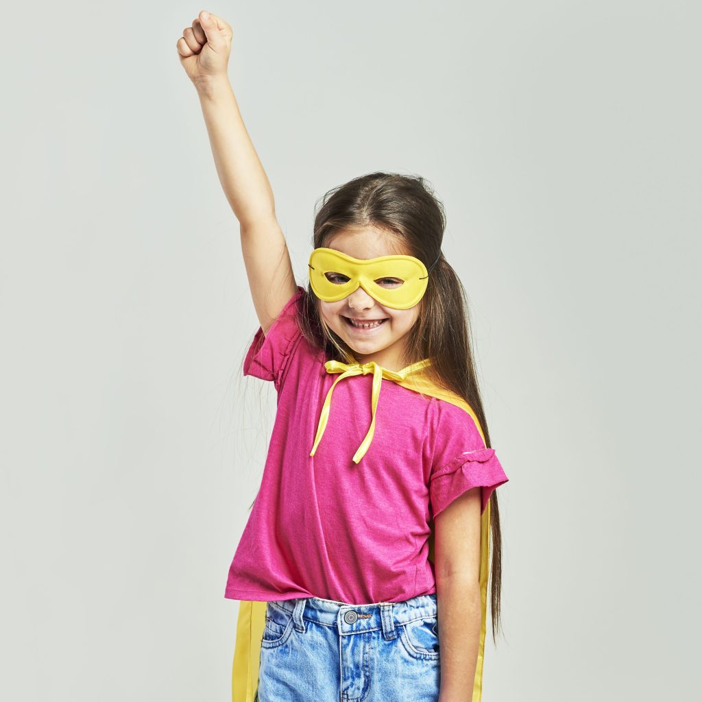 Mädchen zwischen 6 und 8 Jahren mit pinkem T-Shirt, Jeans, einem gelben Cape und gelber Superheldin-Maske steht frontal lächelnd und hebt ihre rechte Faust hoch wie eine Superheldin. Sie symbolisiert die Mädchenkurse für Selbstbehauptung für 6- bis 8-Jährige.